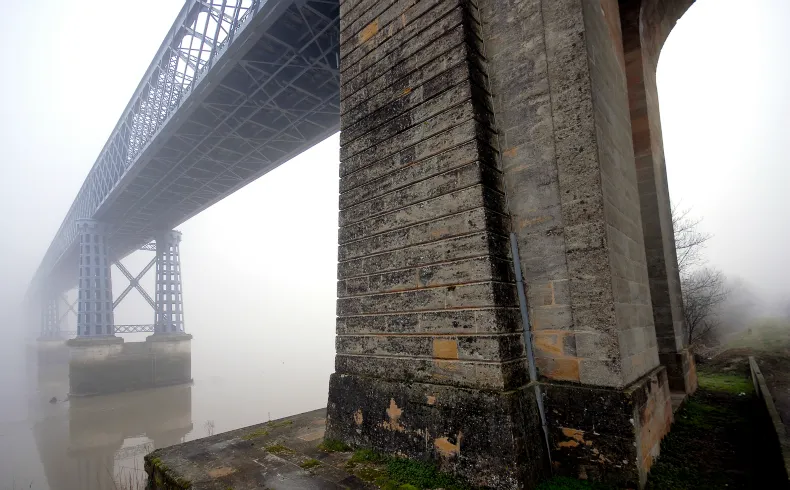 Misty view beneath Bordeaux's stone bridge showing massive stone pillars and the metal structure of the Eiffel bridge in the background, with green algae on the foundations.