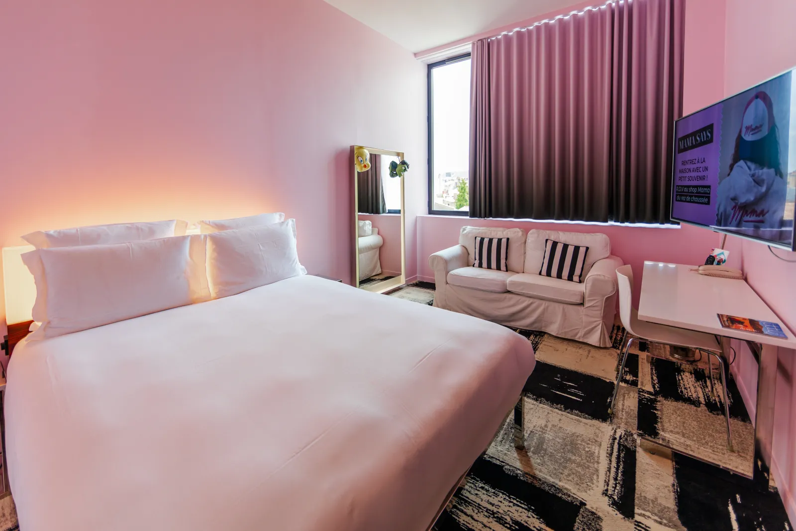 Modern bedroom at Mama Shelter Bordeaux featuring pink walls, white bed, beige sofa, wall-mounted TV, and large window with curtains.