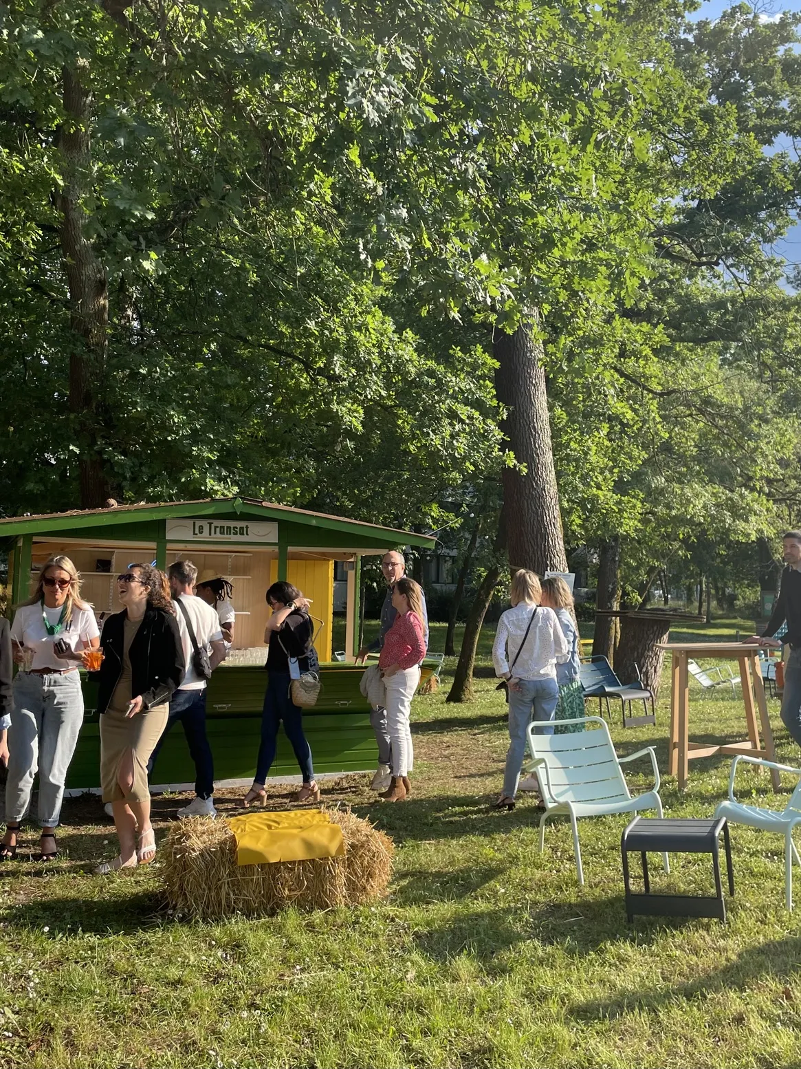 The Transat event in Bordeaux featuring a green and yellow food stand, visitors gathered in a shaded park with large trees, blue deck chairs and street furniture.