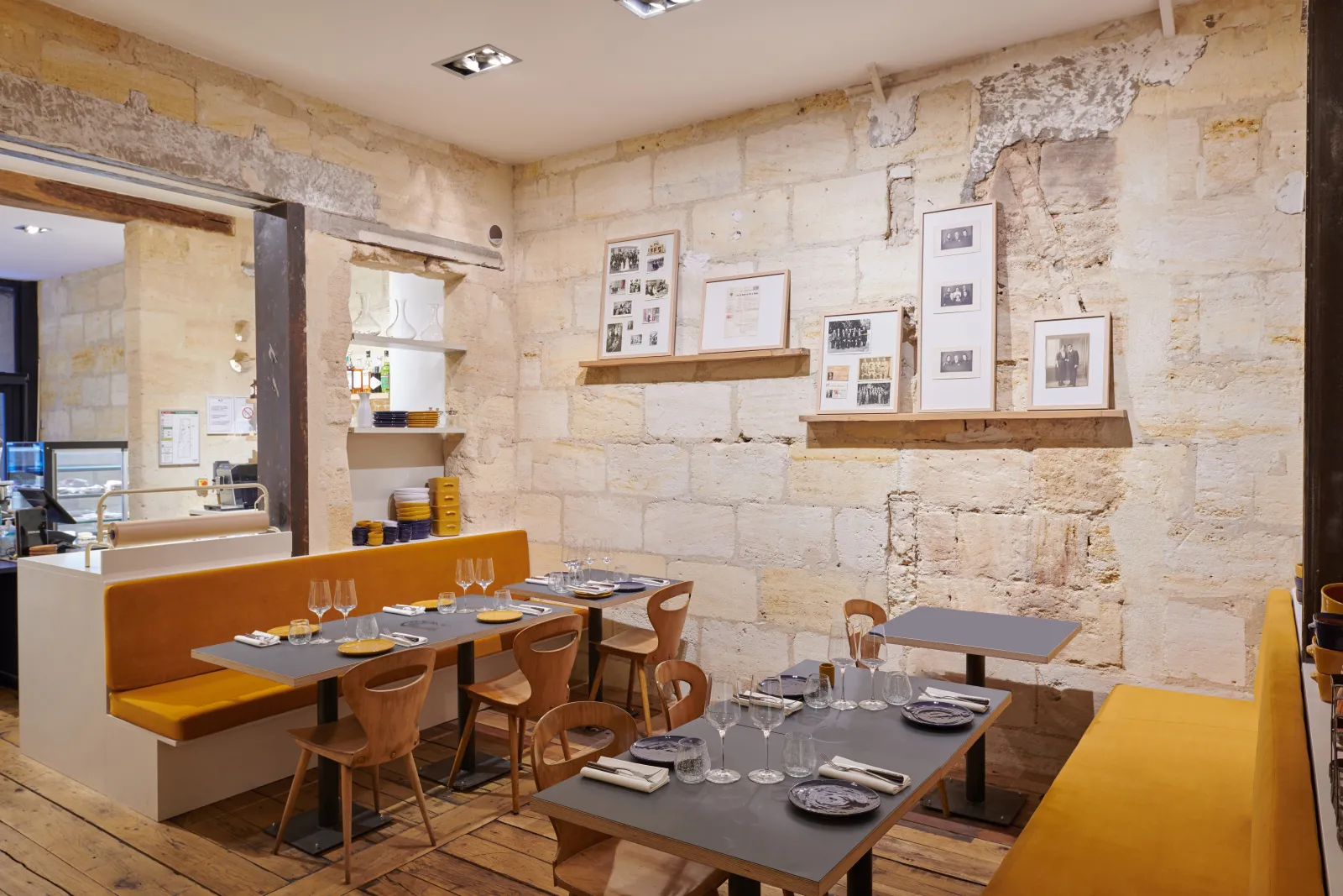 Contemporary restaurant interior in Bordeaux featuring exposed stone walls, grey dining tables, yellow and orange upholstered bench seating, light wood chairs, vintage photographs on floating shelves, and a modern counter area.