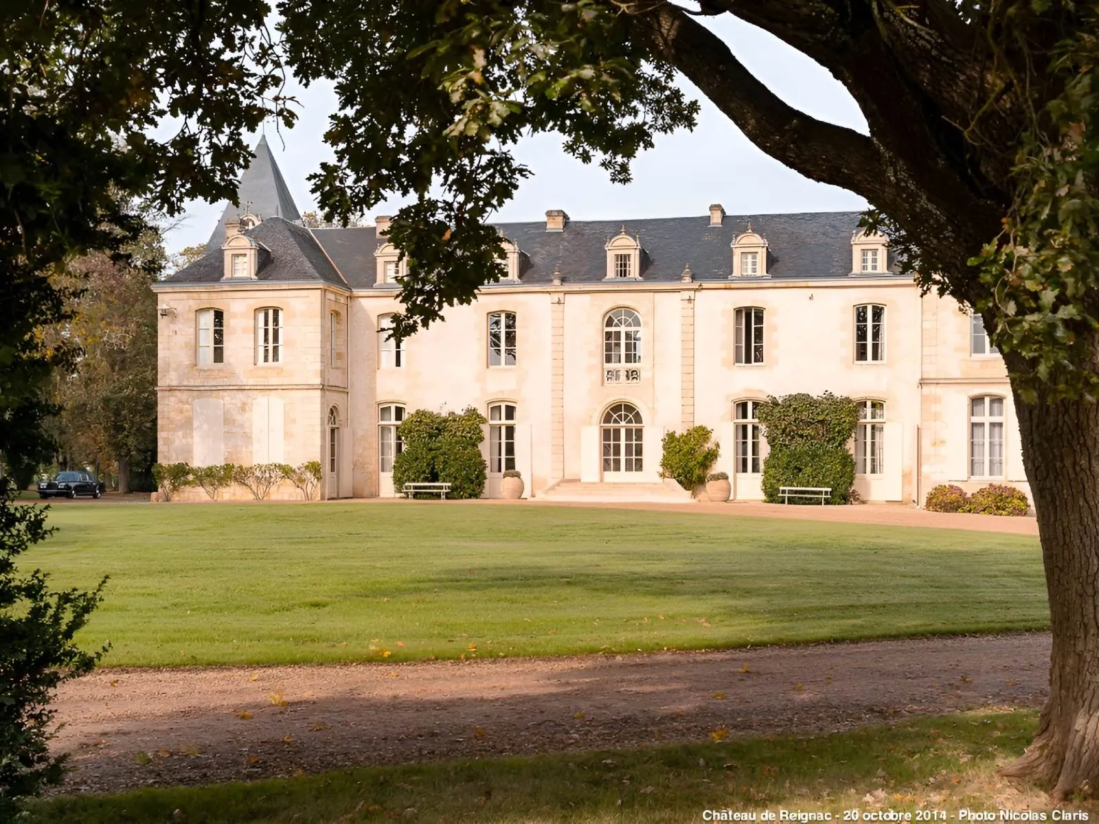 The Château de Reignac, an elegant 19th-century stone mansion with a mansard roof, framed by tall trees, overlooking a green lawn and gravel driveway.