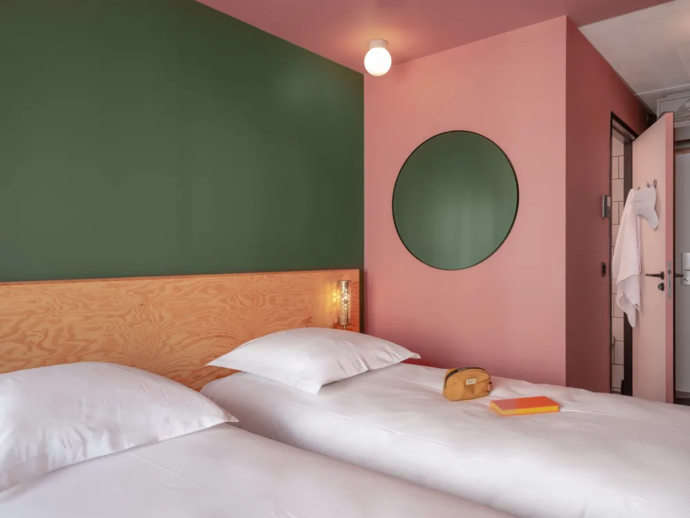 Modern hotel room at Eklo Bordeaux featuring two twin beds with natural wood headboard, color-blocked walls in soft green and pink, round green mirror, and minimalist design.