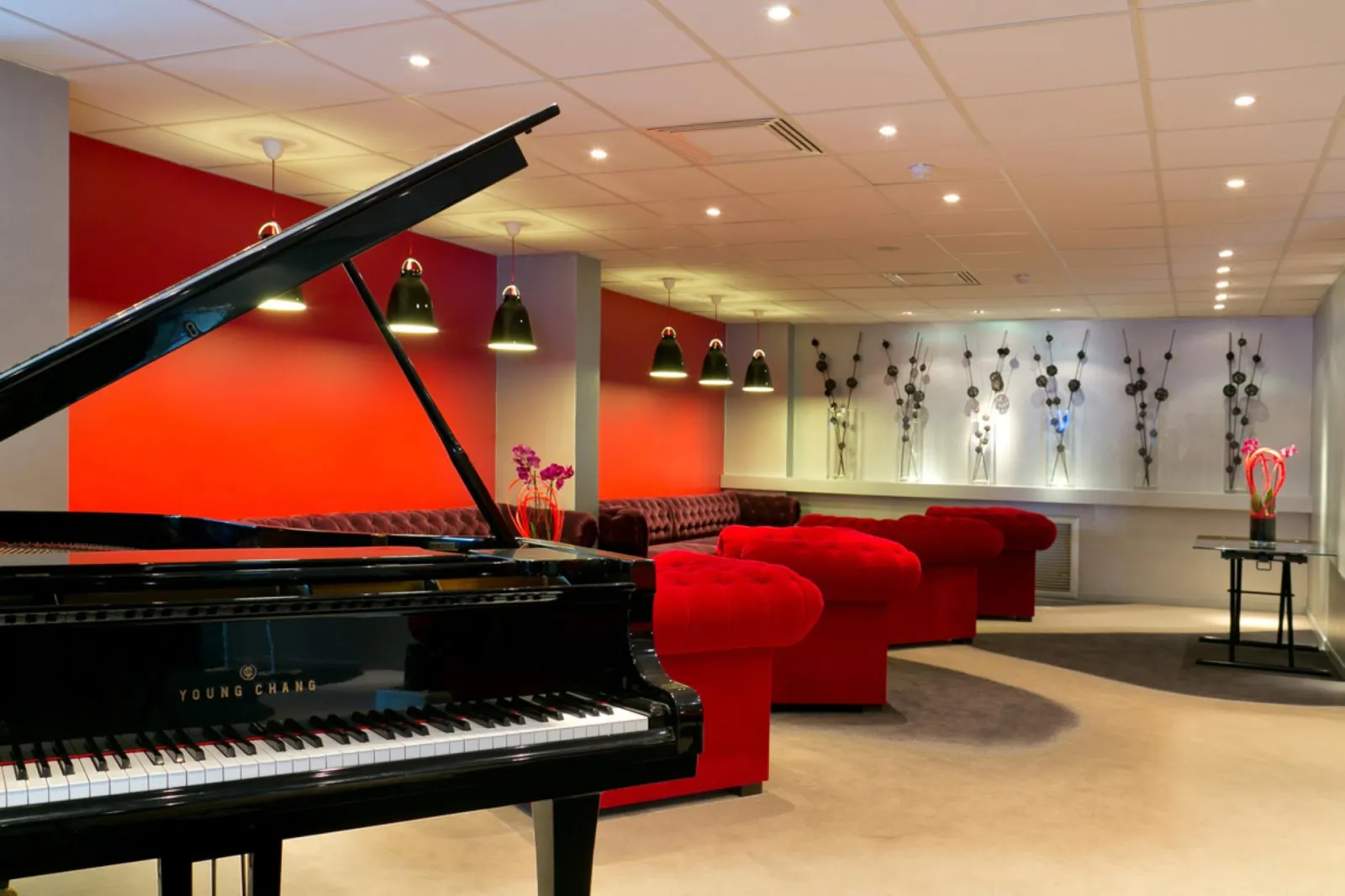 Modern lobby of Mercure Bordeaux-Lac Hotel featuring a black Young Chang grand piano, red designer armchairs, red and beige walls, recessed ceiling lights, and hanging floral decorations.
