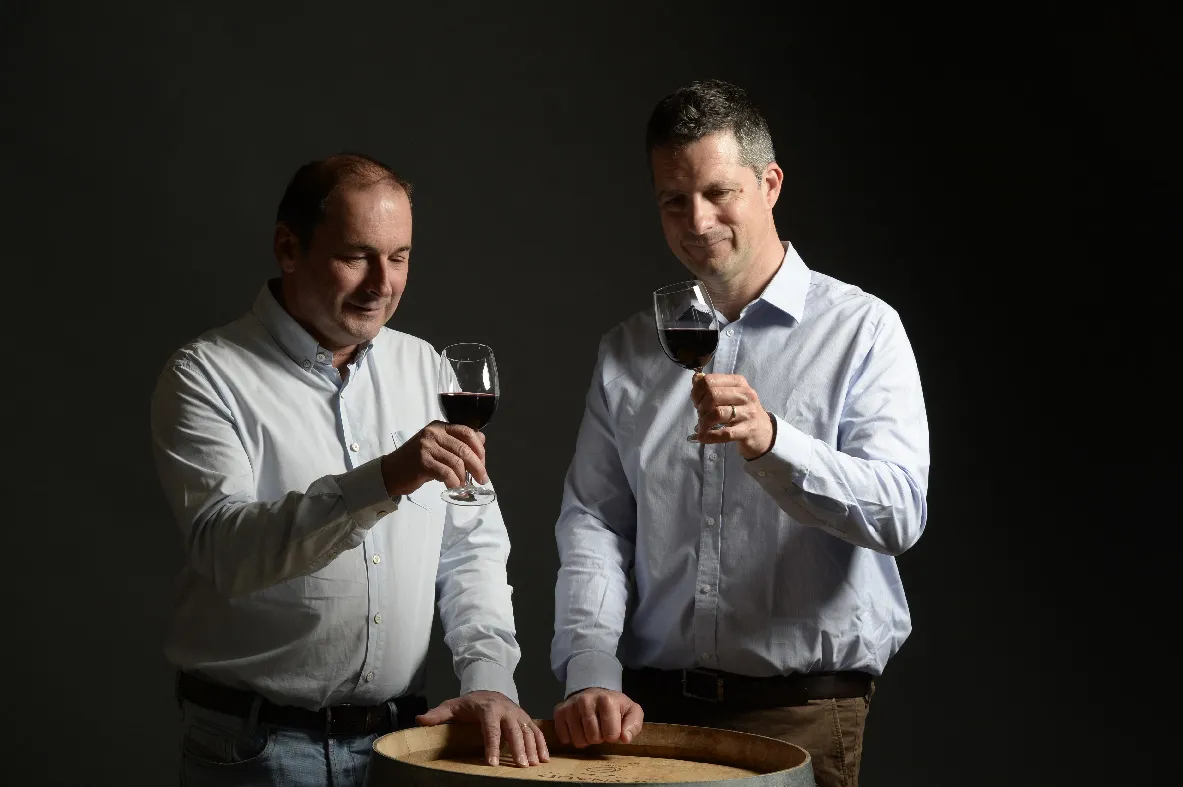 Two men in white shirts toasting with glasses of red wine in front of a wooden barrel in the cellars of Château Lagrange in Bordeaux.