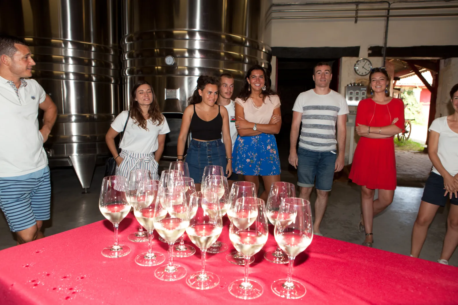 Group of visitors standing around a table with white wine glasses in front of a stainless steel fermentation tank at the Ateliers au Château in Bordeaux.
