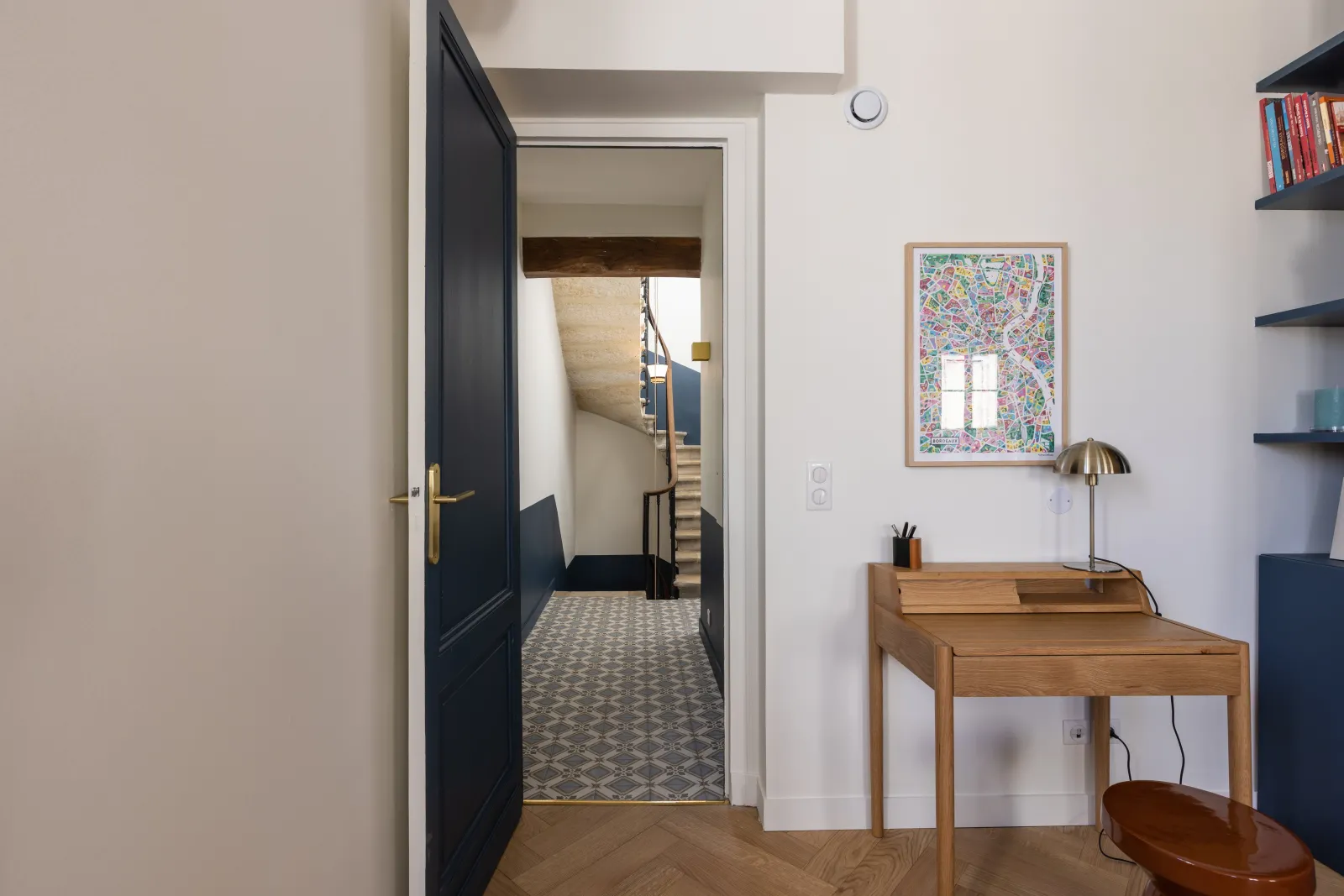 Modern home office interior in Bordeaux featuring a light wood desk with a brass lamp, floating shelves, an open black door leading to a tiled hallway, and wall décor elements