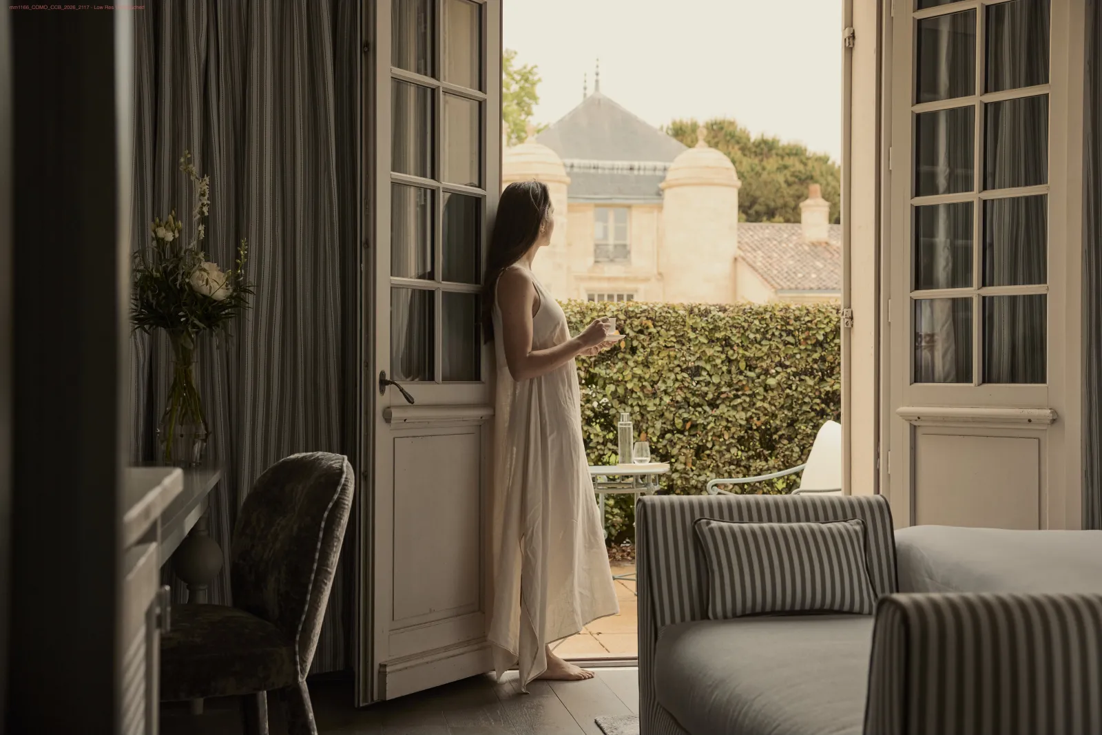 Woman in a white dress looking out through open French doors from an elegant bedroom overlooking a manicured garden and château in the distance at Bordeaux.