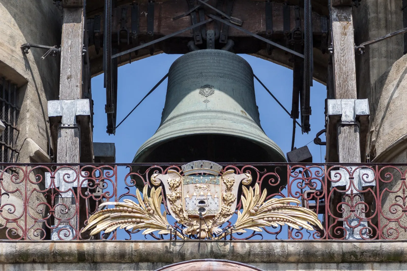The Great Bell of Bordeaux viewed from below, displaying a large bronze bell suspended in a wooden frame, with an ornate gilded iron balcony and coat of arms beneath, against a clear blue sky.