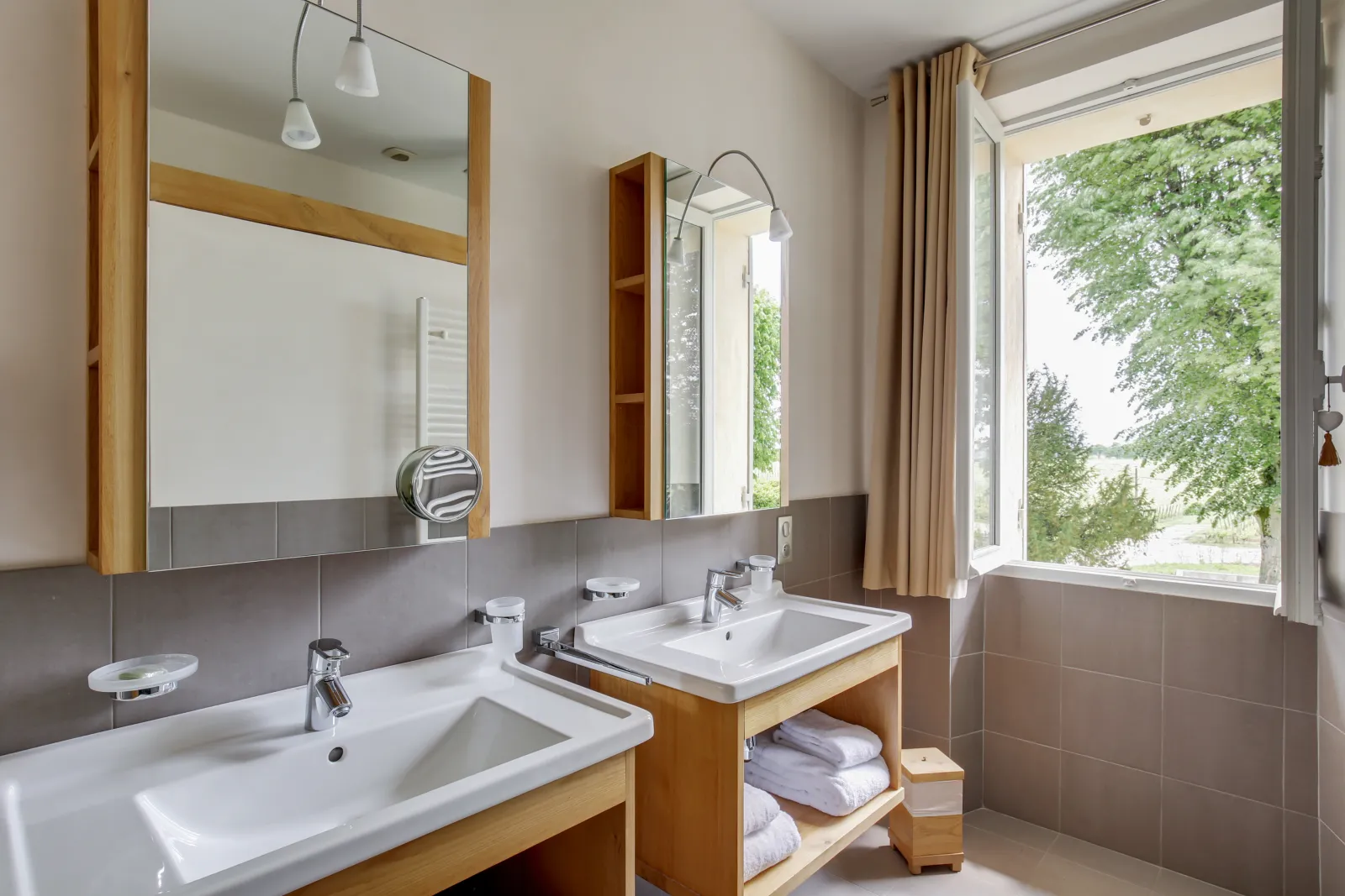 Modern bathroom at Logis de Valandraud in Bordeaux featuring dual white sinks, wooden-framed mirrors, grey and white tilework, bathtub, natural wood storage, and a large window overlooking the garden.