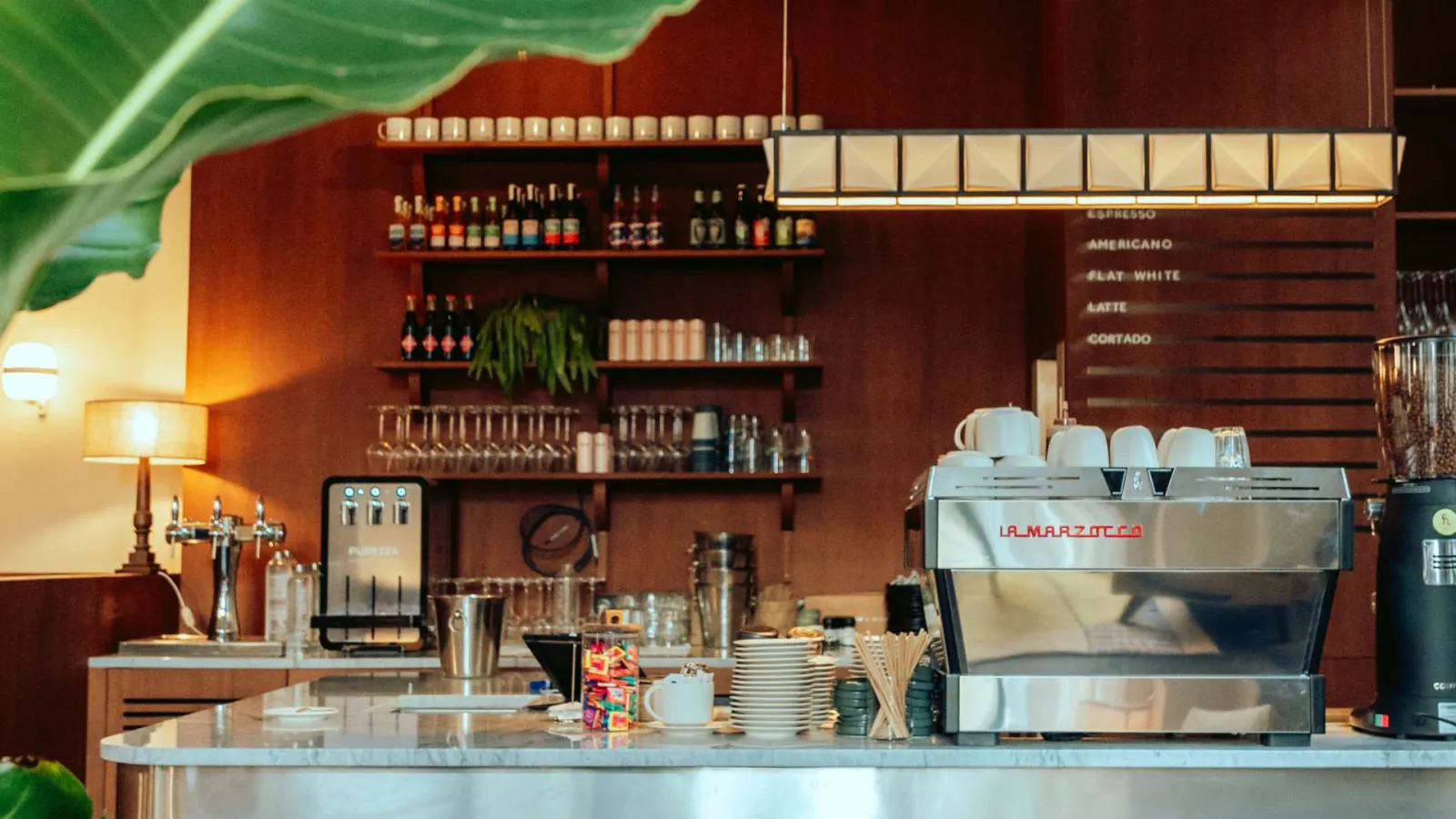 Coffee shop counter in Bordeaux featuring a La Marzocca espresso machine, bottle shelves, wall menu, vintage lamp and green plants with warm-toned décor.