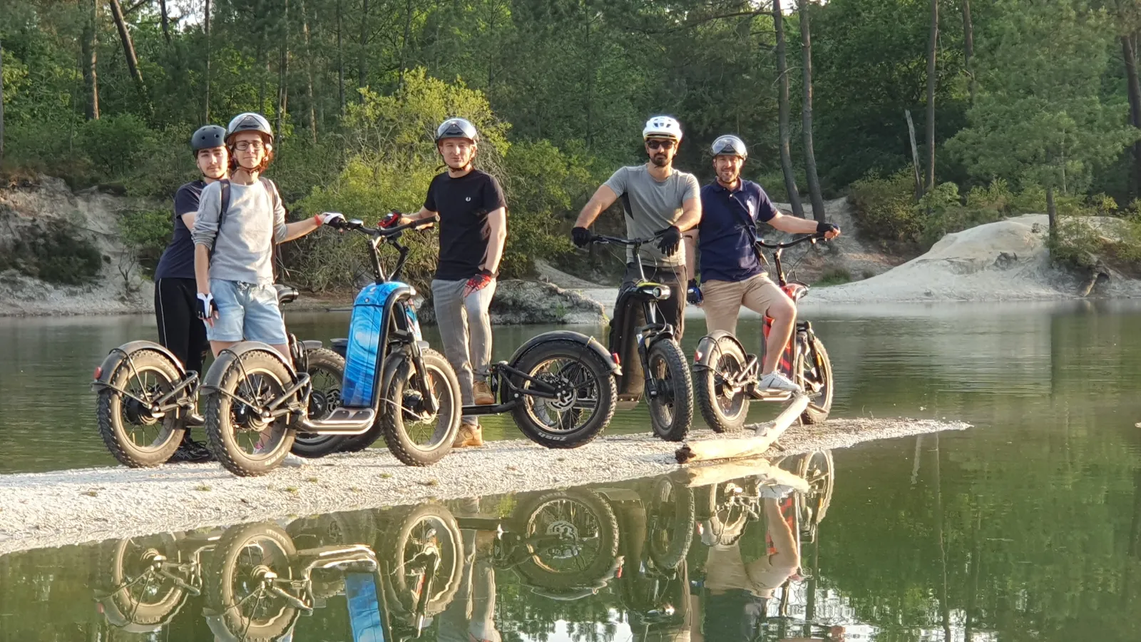 Five people wearing helmets pose with their bicycles and electric scooters on a pebble beach by a river, with a forest in the background.