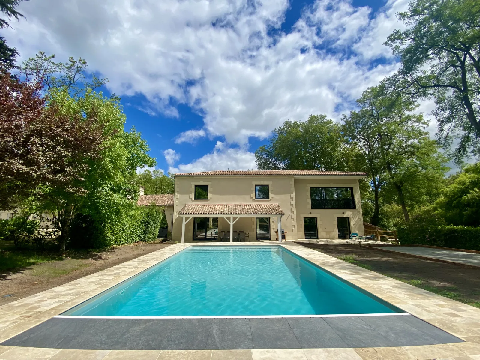 Modern villa with rectangular swimming pool surrounded by lawn and trees, beige facade with covered terrace, cloudy blue sky.
