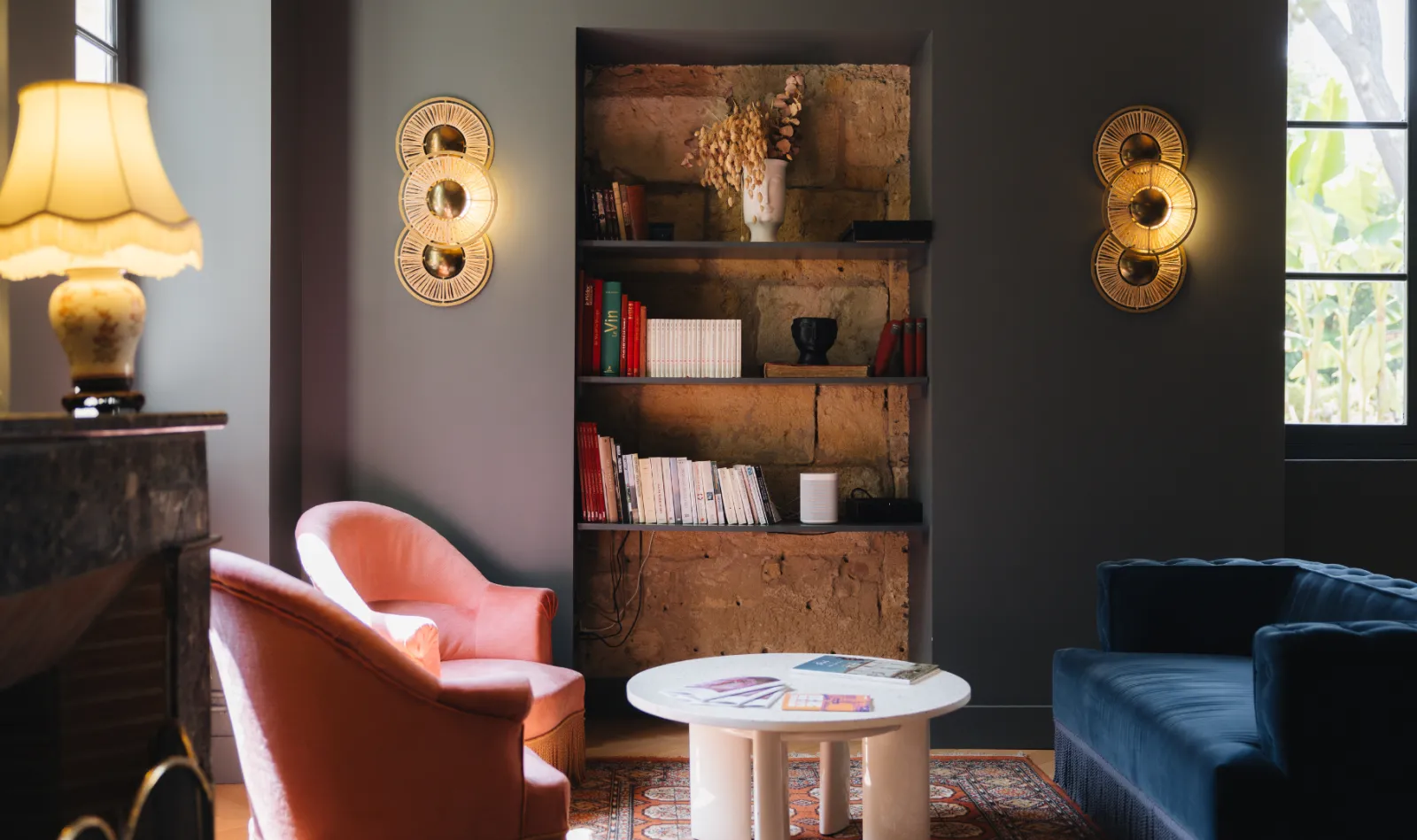 Elegant living room at Château Réal in Bordeaux featuring a coral armchair, rustic shelving, bookcase, golden wall sconces, and navy blue furniture.