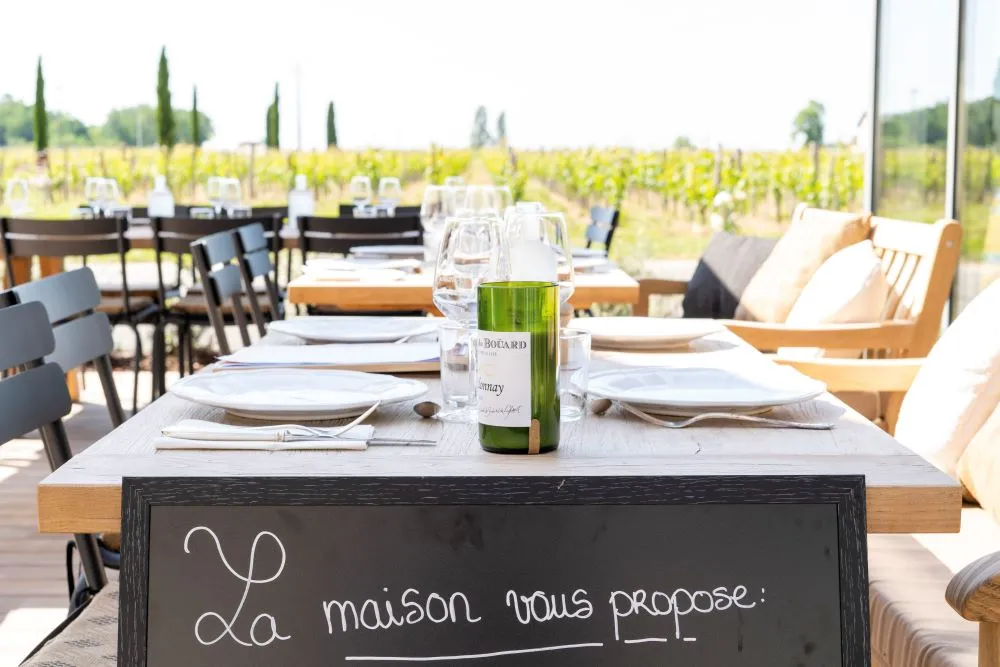 Outdoor dining table overlooking Bordeaux vineyards with Boüard wine bottle, place settings, and chalkboard sign reading 'La maison vous propose'