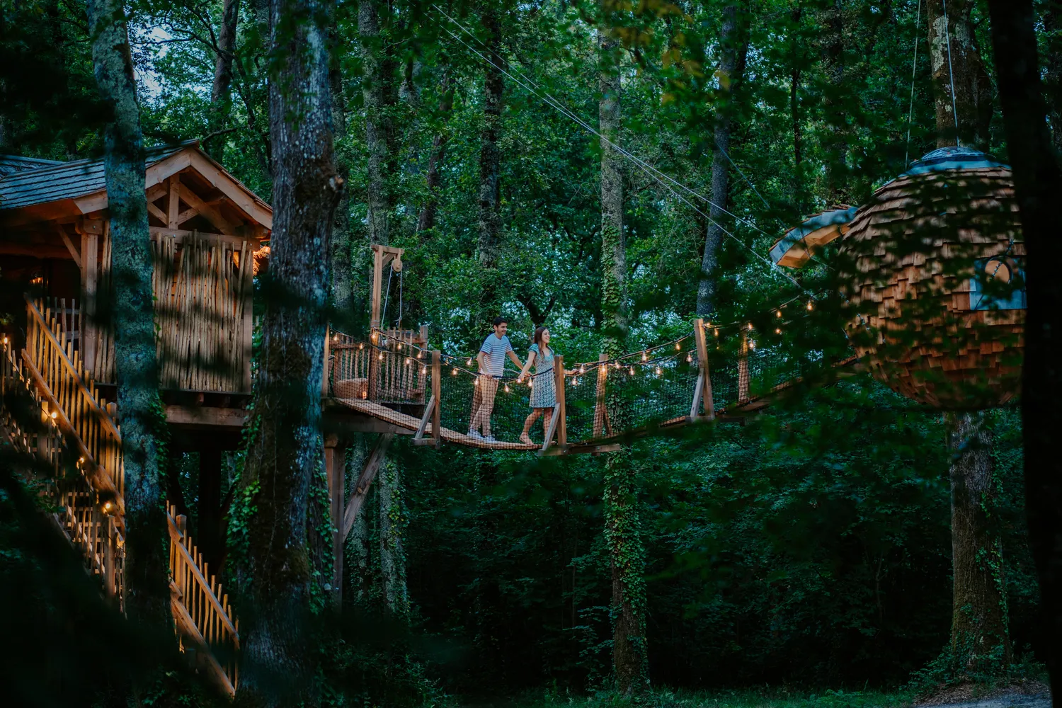 Two people walk on a suspended walkway between wooden treehouses lit by string lights nestled in a dense forest.