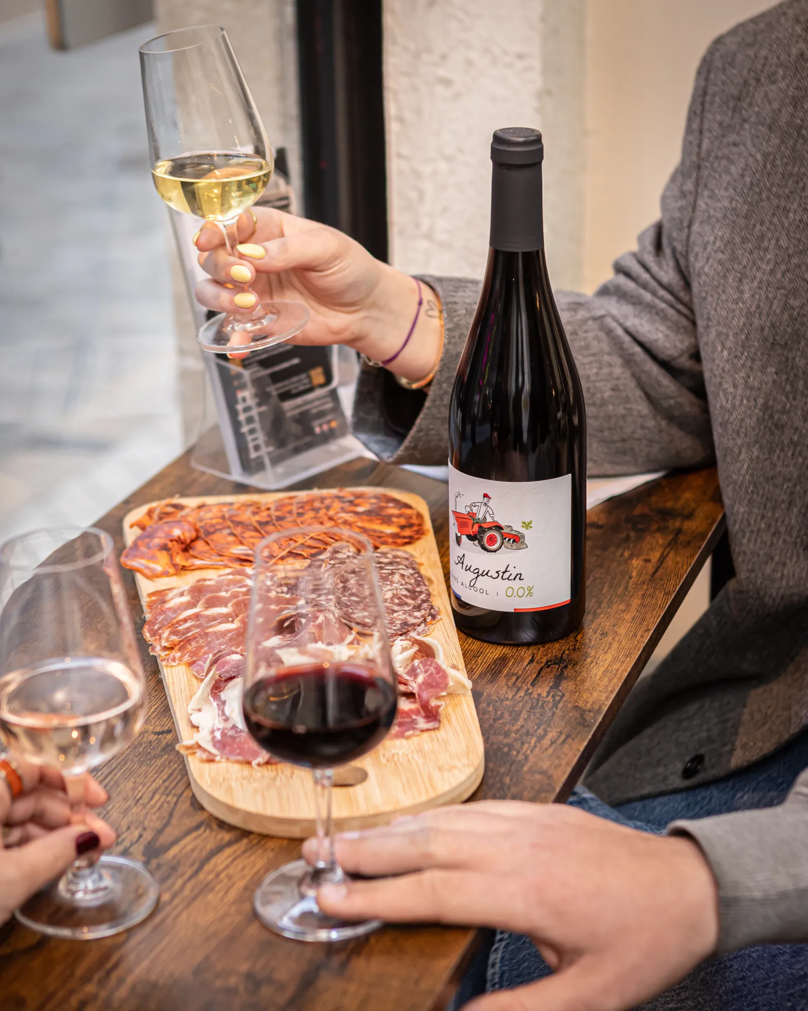 Two people enjoy white and red wine paired with a charcuterie and cheese board, with a red wine bottle labeled Augustin displayed on a wooden table.