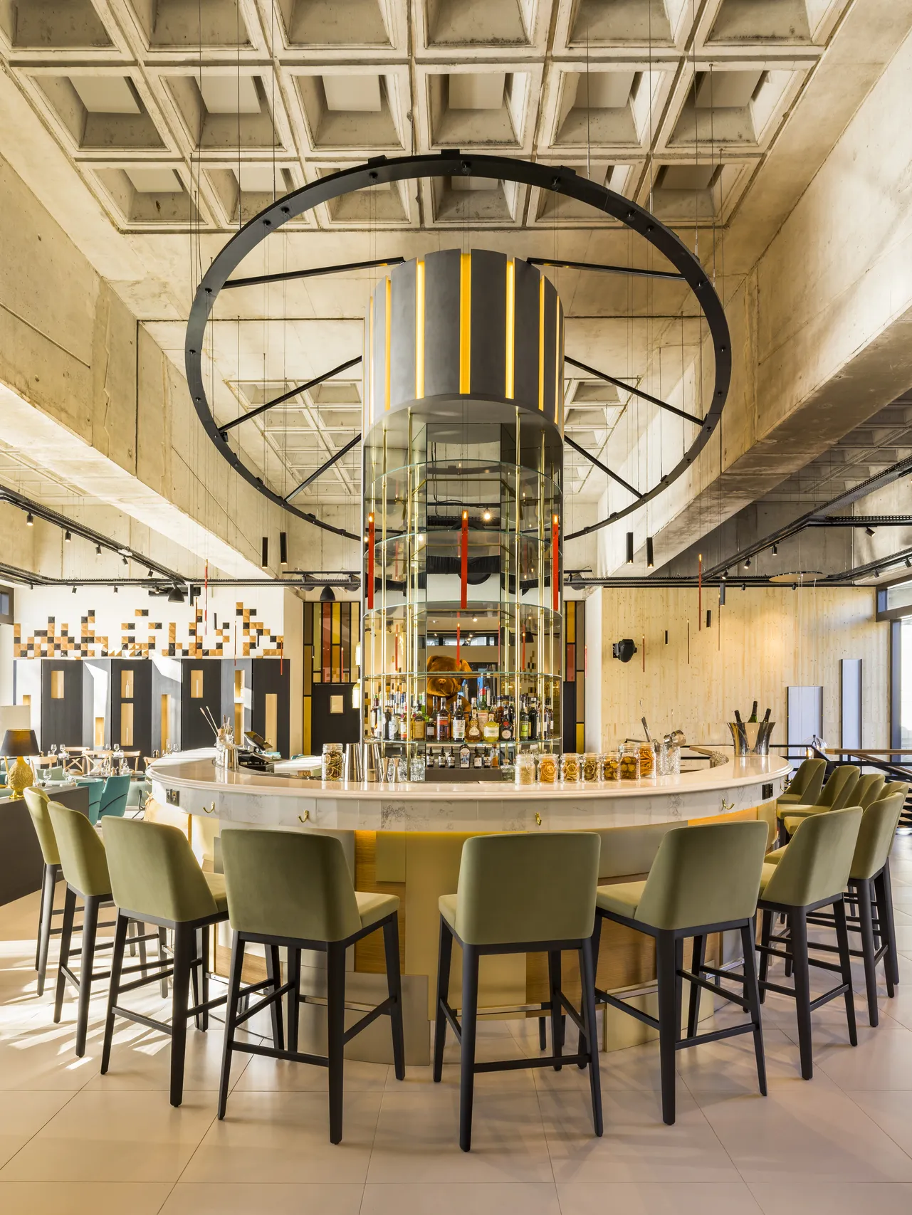 Interior view of Bada in Bordeaux featuring a modern circular bar with green chairs, topped with a large circular metal structure and a geometric coffered concrete ceiling.