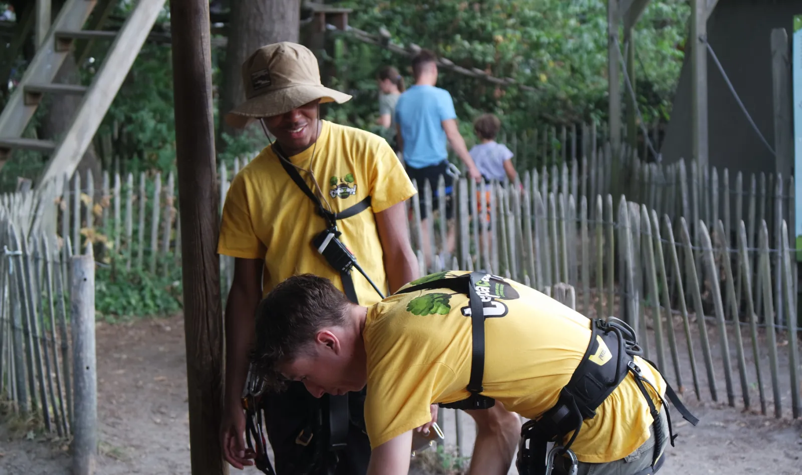 Two instructors in yellow uniforms help a child put on safety harnesses at a treetop adventure park, with other visitors in the background.