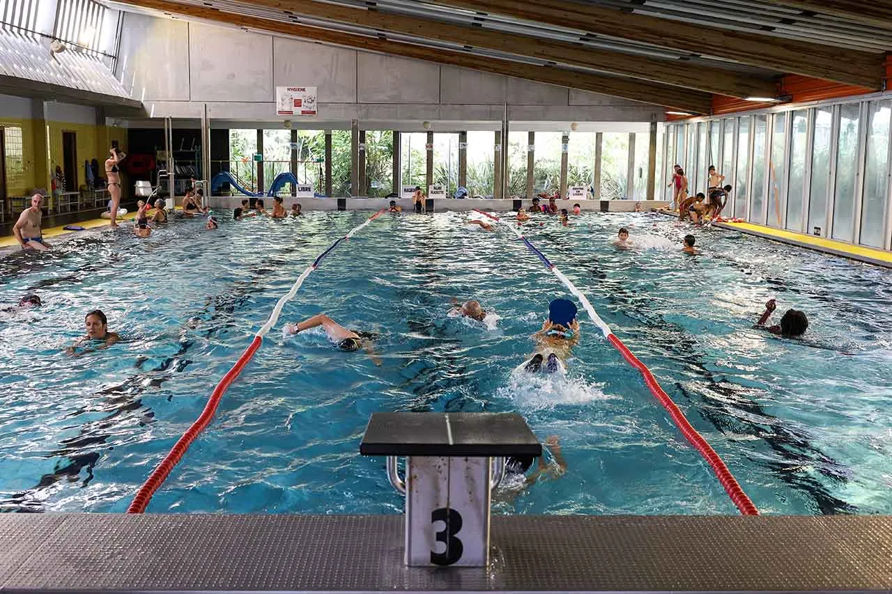 Interior view of the Les Bains municipal swimming pool in Bordeaux with multiple swimmers in lanes separated by red lines, other bathers in the background, and glass windows overlooking an outdoor area.