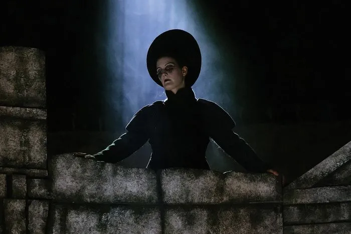 Actress in black costume standing on a stone rampart, lit by dramatic blue lighting, in a production of Dido and Aeneas at Bordeaux Opera.