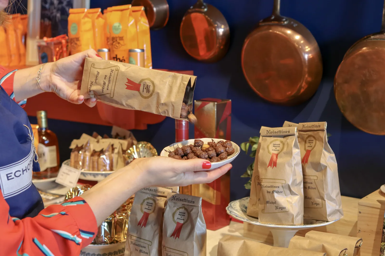 Display of artisanal products at Echoppe de la Lune in Bordeaux: a person holds a bag of hazelnuts and a plate of chocolates, surrounded by packages of noisettines and copper decorative objects.