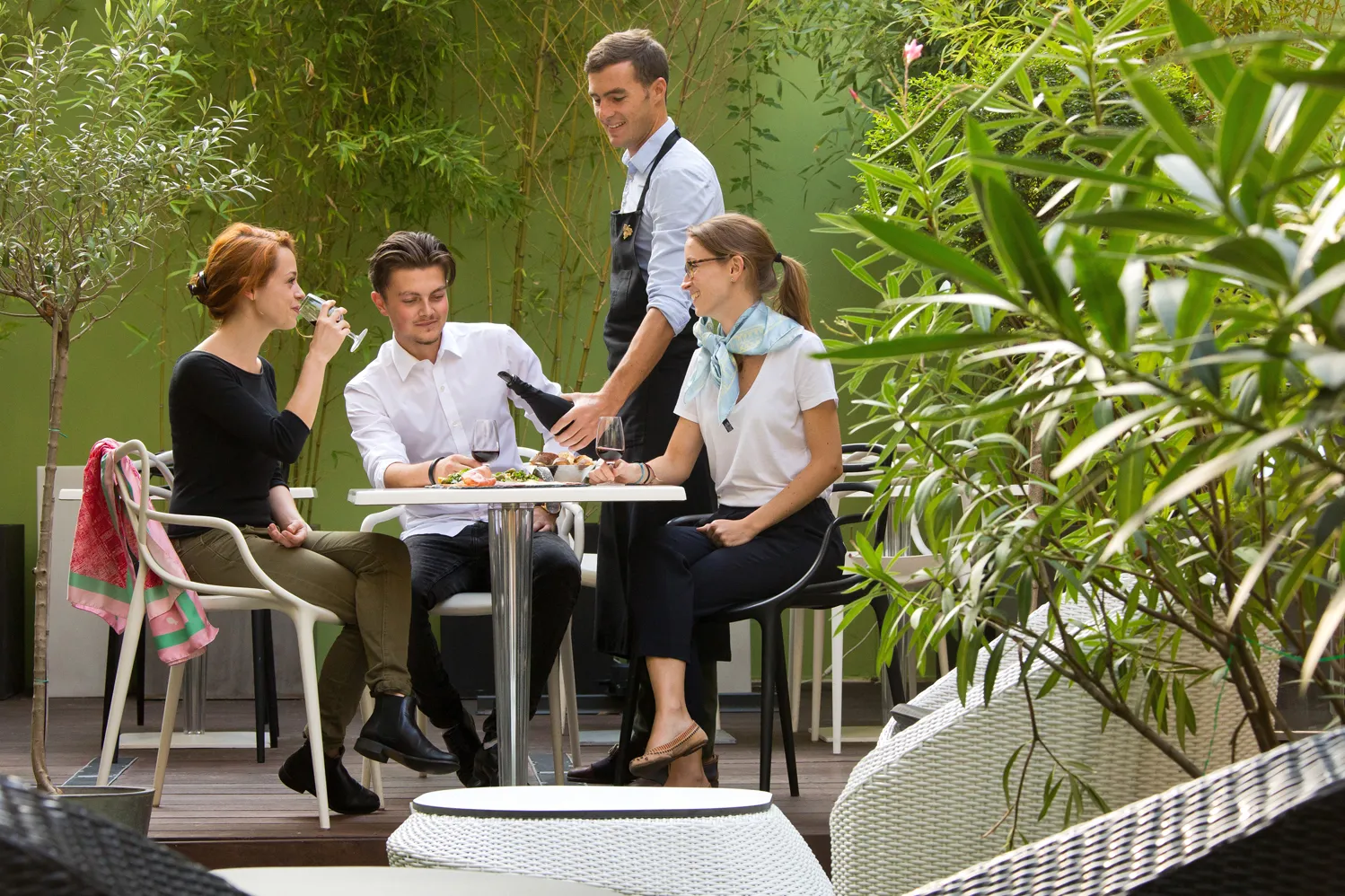 Two guests enjoy a meal at a high table in the lush garden terrace of Bordeaux's Boutique Hotel Bar, while a waiter presents a wine bottle.