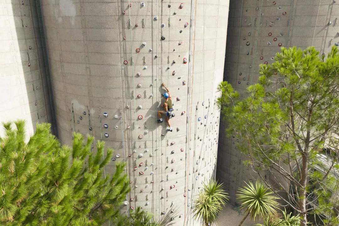 A climber ascends a concrete climbing wall with colored holds at Moxy Bordeaux hotel, surrounded by green vegetation.