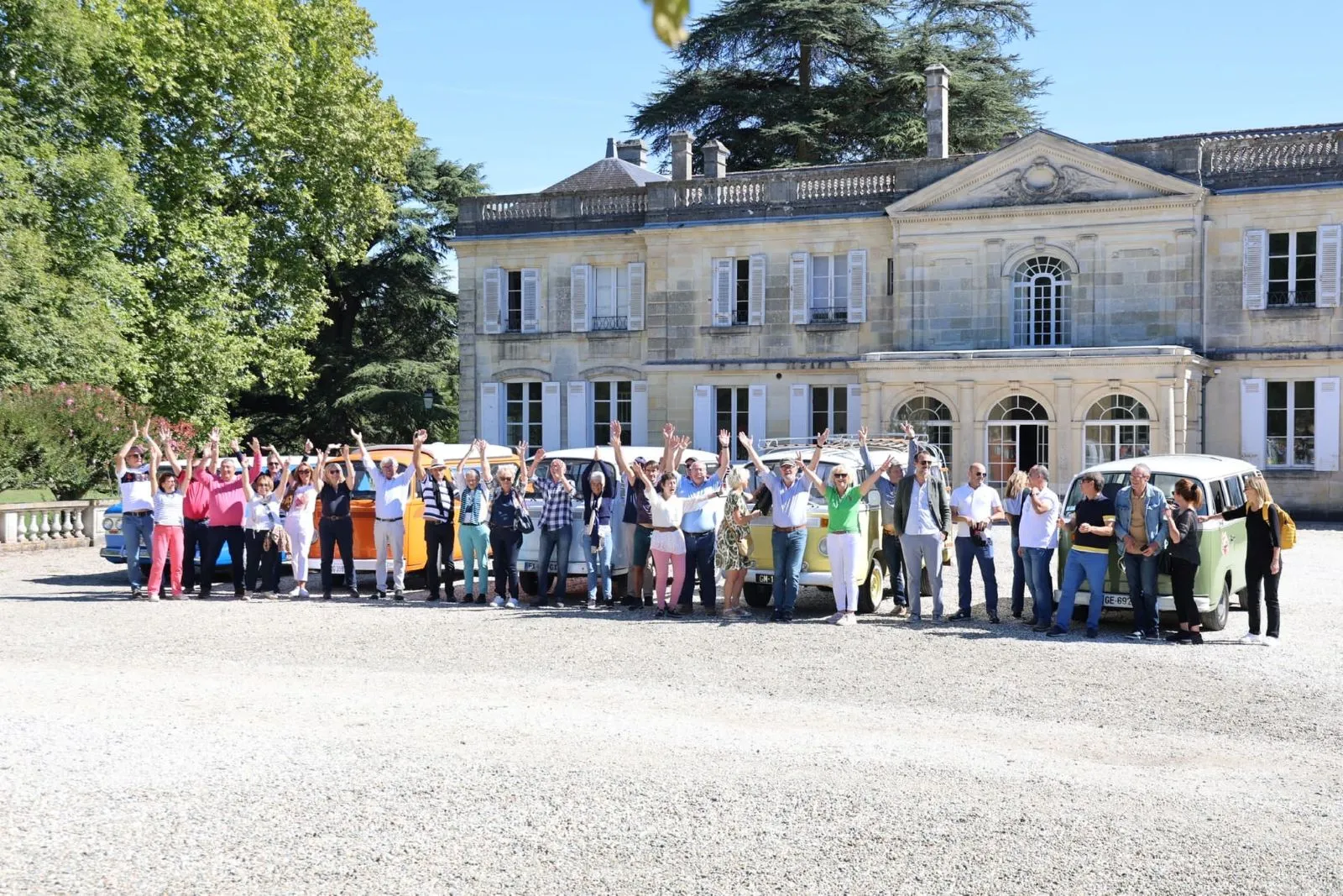 Group of tourists posing in front of colorful vintage camper vans in front of a classical mansion in Bordeaux on a sunny day.