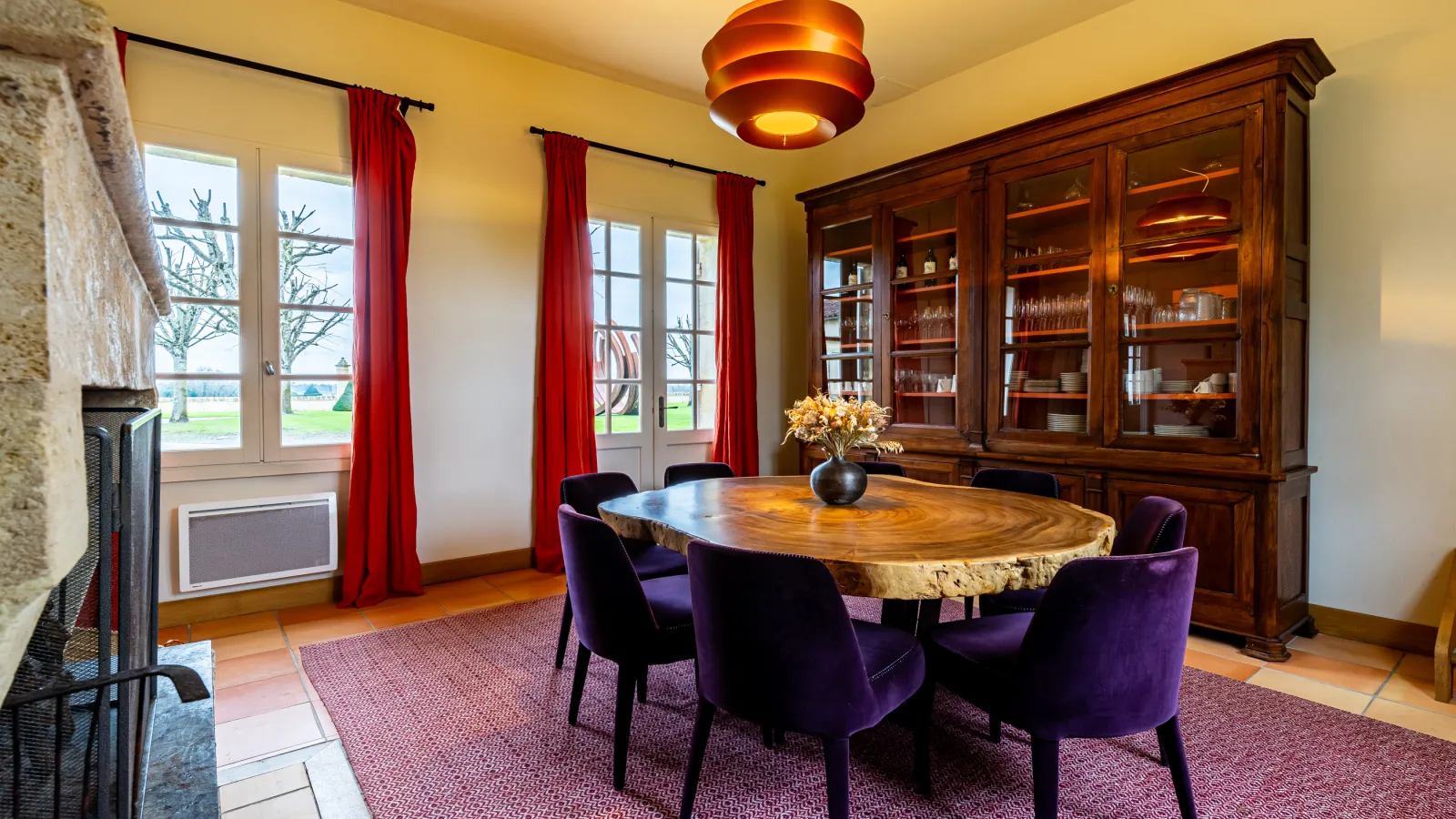 Elegant dining room at Château Malescasse featuring a round natural wood table, purple upholstered chairs, wooden glass-fronted cabinet, French windows with red curtains overlooking the grounds, gold ceiling light, and fireplace.