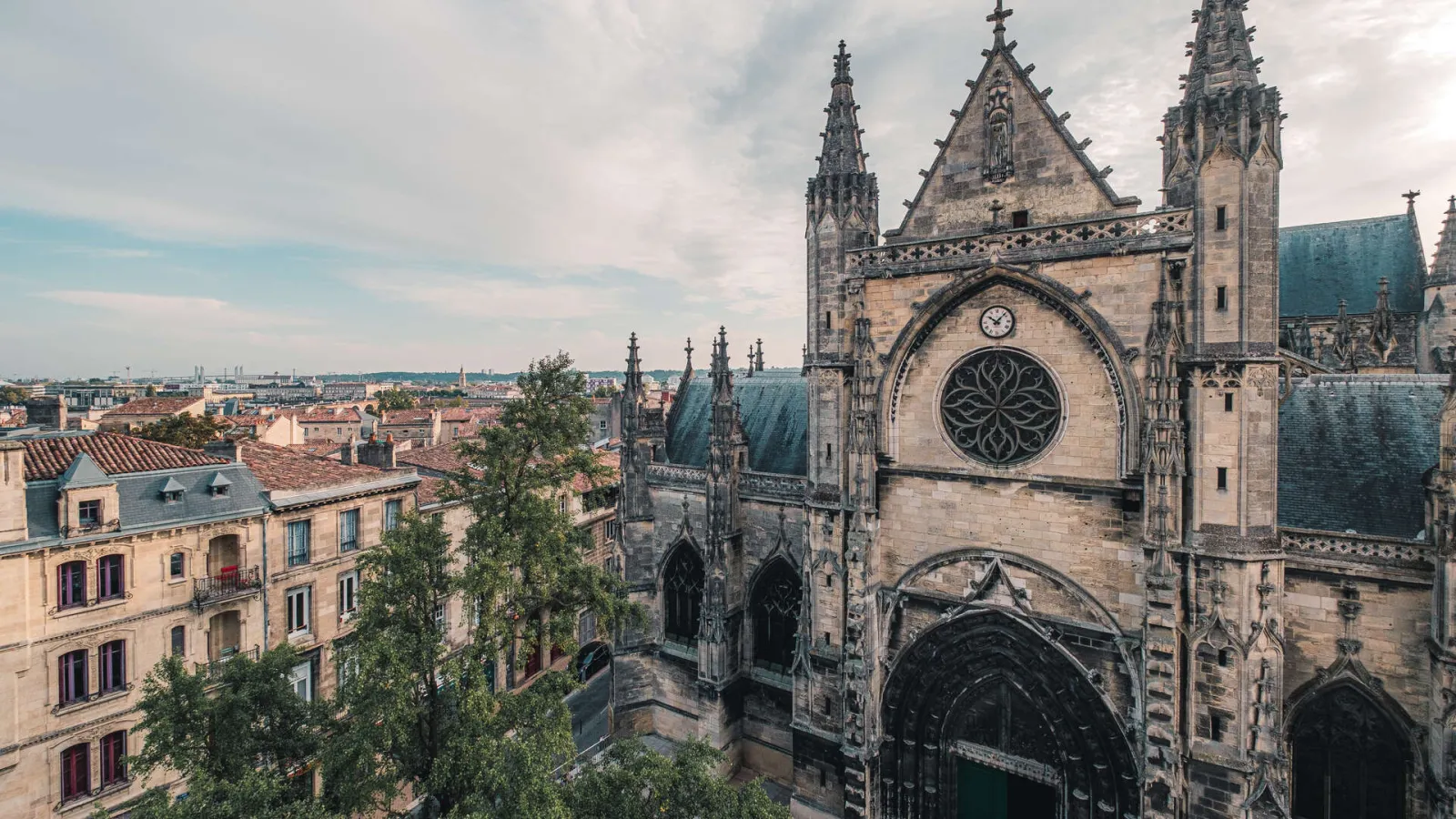 Aerial view of the Basilica of Saint-Michel in Bordeaux featuring Gothic spires, rose window, and green slate roof overlooking the historic city's terracotta rooftops.