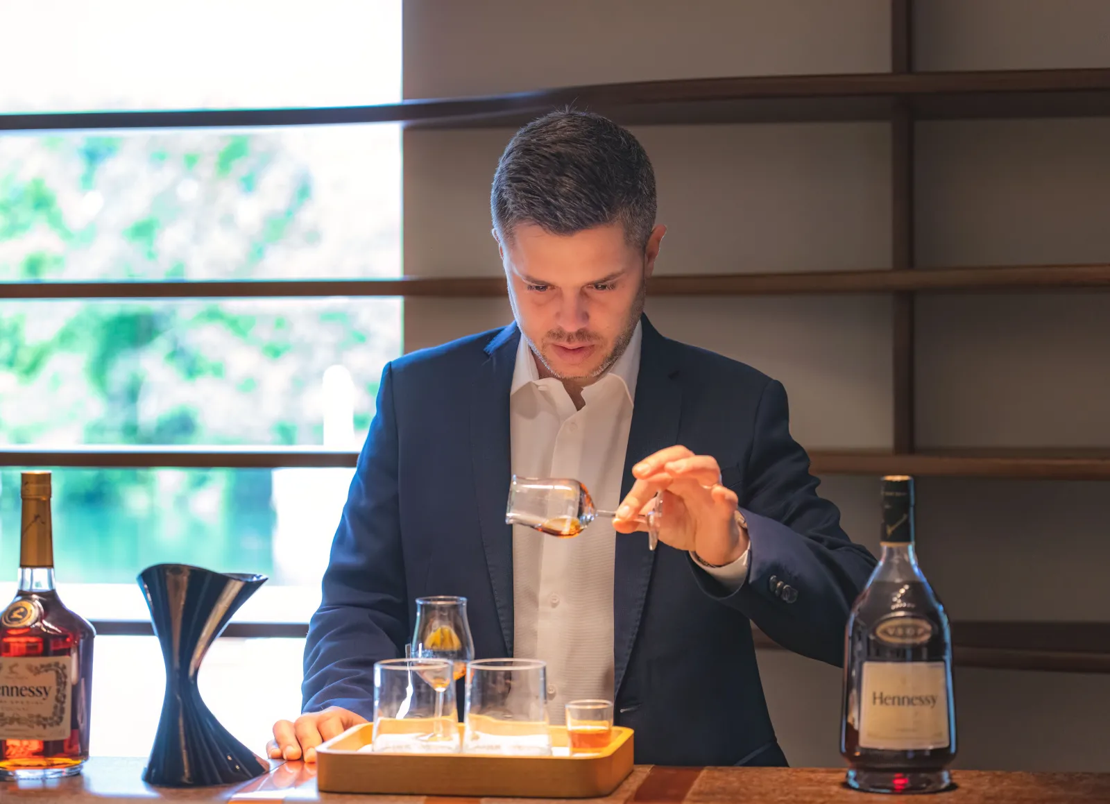 A man in a blue suit presents a Hennessy cognac tasting in an elegant room with bottles and glasses.