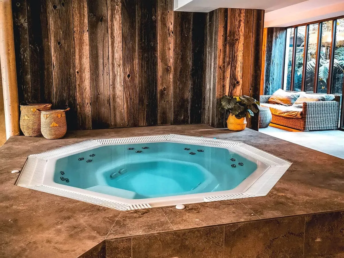 Indoor spa featuring a turquoise hot tub with massage jets, surrounded by rustic wooden walls, woven baskets, and warm-toned relaxation furniture.