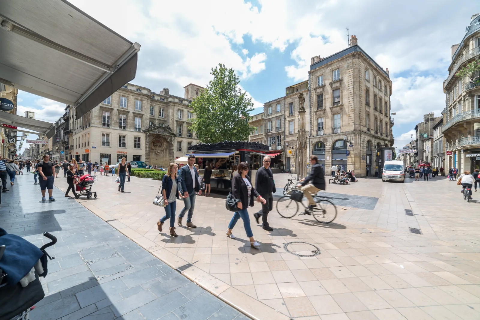 Busy square in Bordeaux with pedestrians, cyclists and shops, lined with Haussmann-style buildings under a cloudy sky.