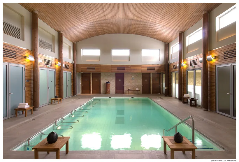 Indoor swimming pool at Les Bains de Margaux spa in Bordeaux, featuring turquoise water, wooden vaulted ceiling, changing rooms, relaxation benches, and ambient lighting.