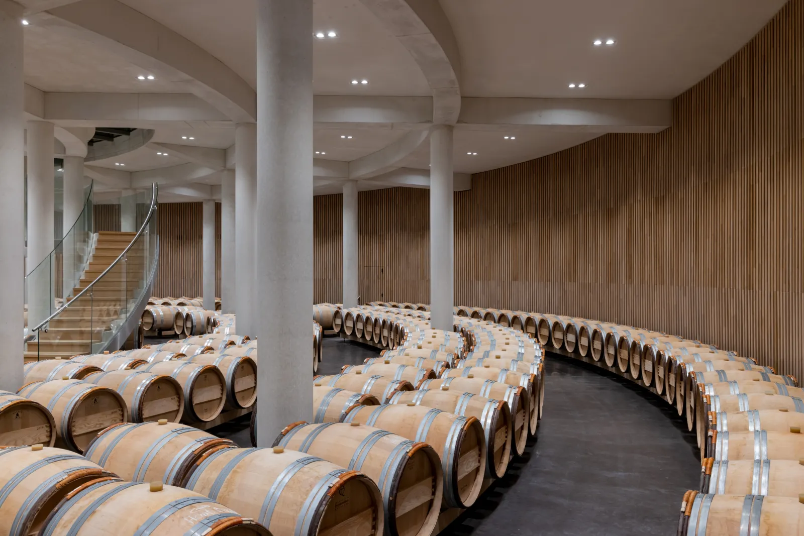 Modern winery cellar at Château Haut-Bailly in Bordeaux featuring symmetrical rows of oak barrels, contemporary ceiling with white beams and supporting columns, subtle LED lighting, wooden and glass staircase visible in the background.