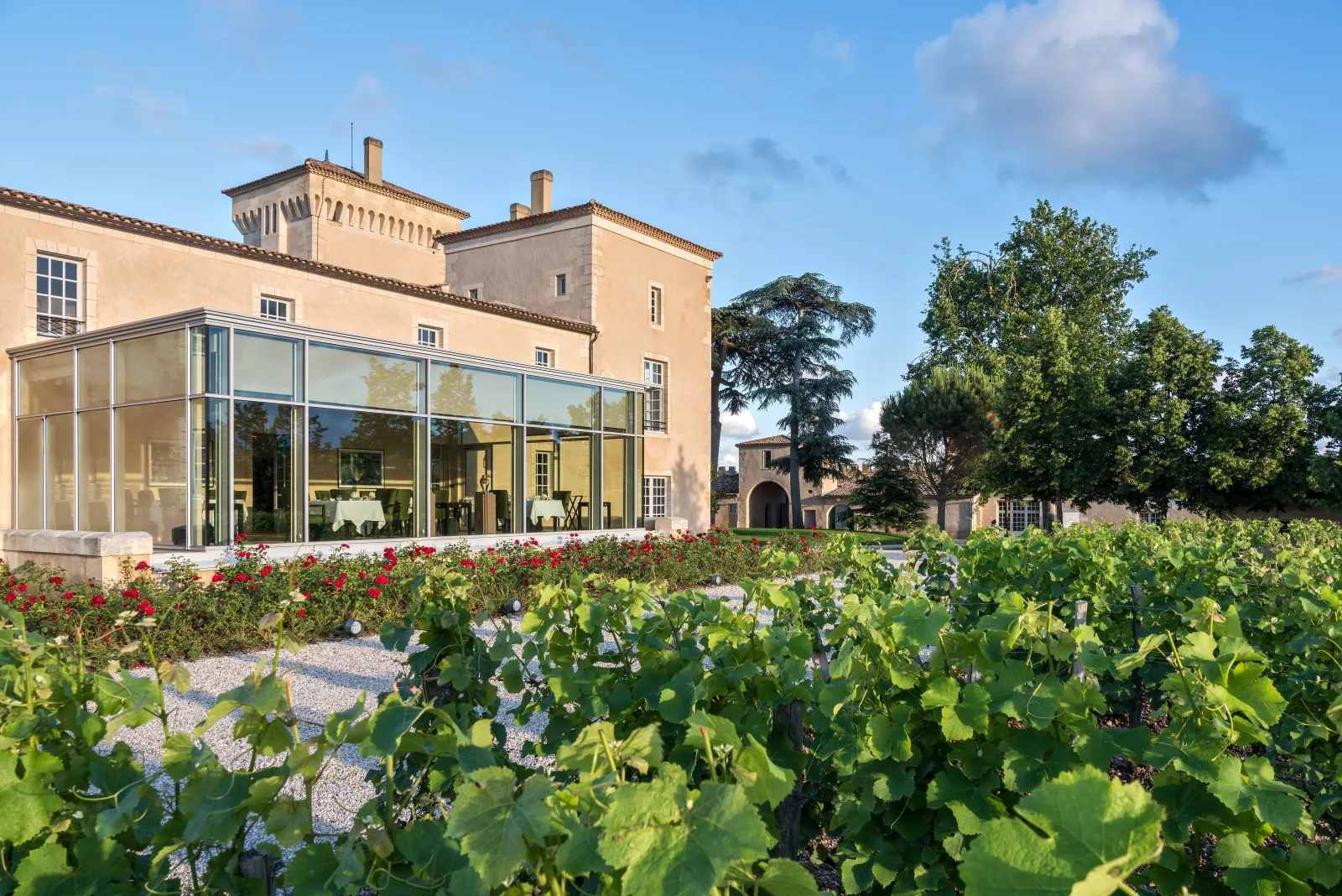 Lalique Hotel at Château Lafaurie Peyraguey in Bordeaux, featuring a modern glass extension attached to a historic ochre-yellow building, with vineyard rows in the foreground and trees under a blue sky.