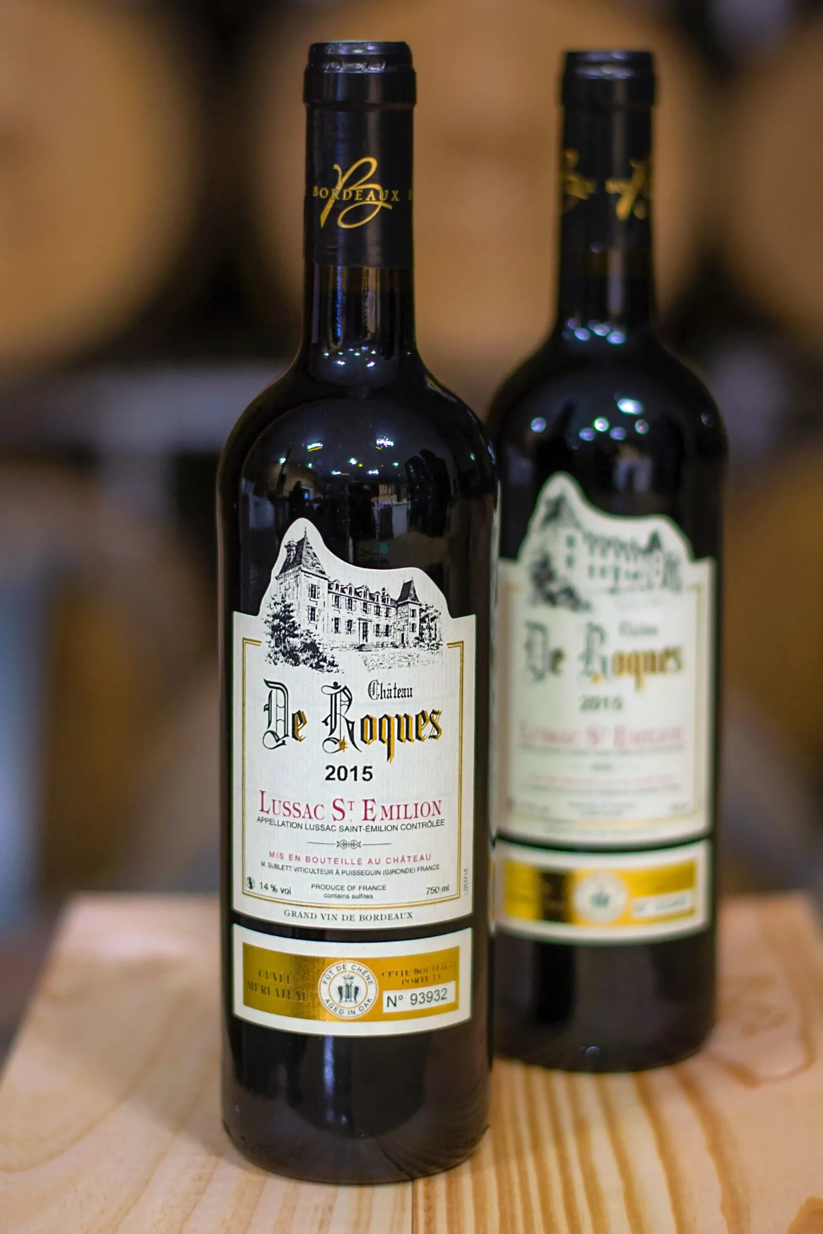 Two bottles of Château de Roques 2015 red wine from Lussac Saint-Émilion, featuring a white label with château imagery and gold band, displayed on a wooden surface.