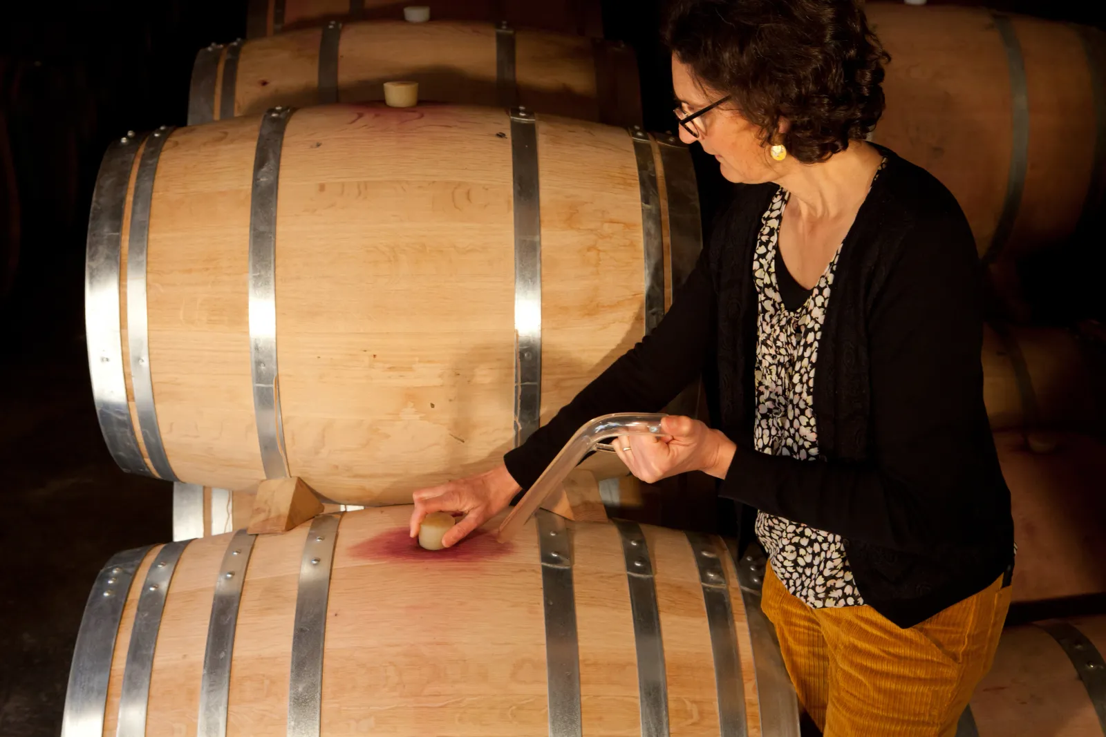 A woman inspects wooden wine barrels in a Bordeaux wine cellar using a quality control instrument.