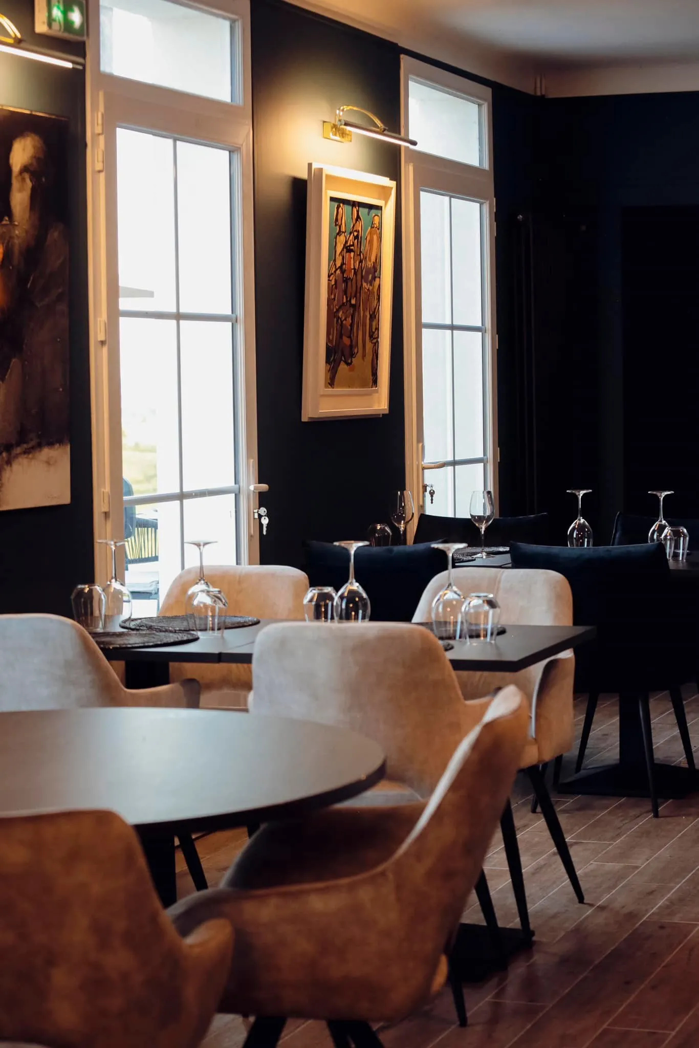 Elegant restaurant dining room in Bordeaux featuring round modern tables, cognac leather chairs, black walls, geometric-patterned tall windows, golden lighting, and classical artwork.