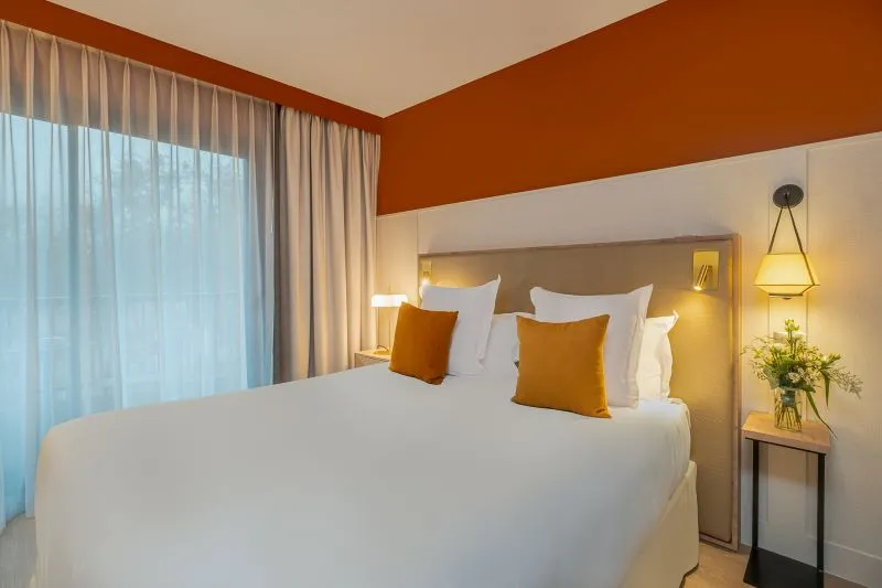 Modern hotel bedroom featuring a white bed with orange pillows, terracotta and beige walls, large windows with beige curtains, bedside table with flowers and pendant light.