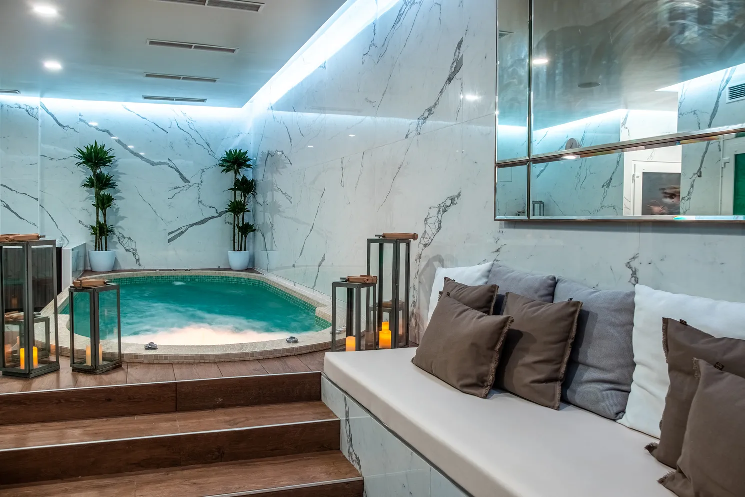 Luxury spa space featuring a turquoise indoor pool, white marble walls, blue LED lighting, relaxation bench with cushions, green plants and wall mirror