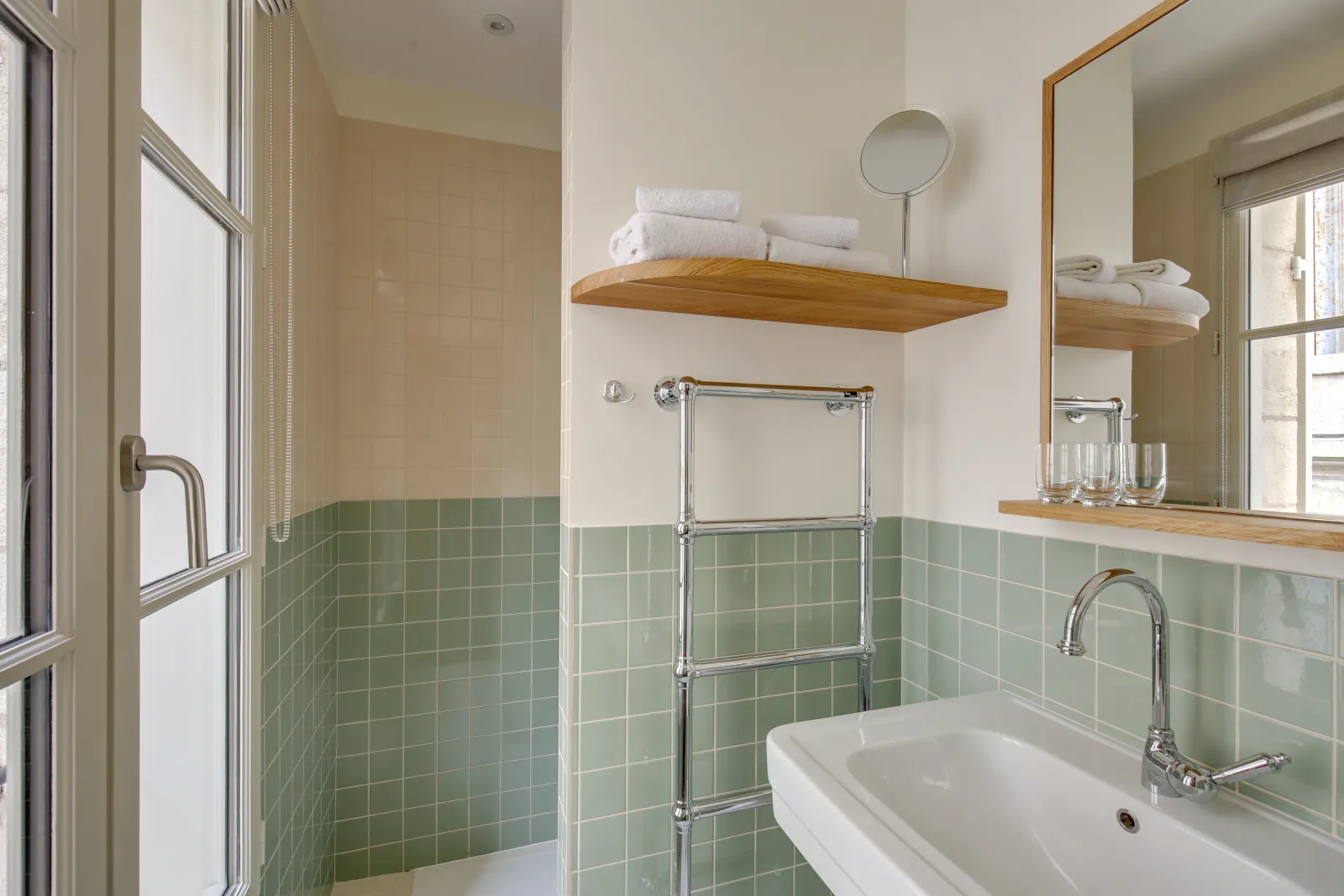 Modern bathroom with light green tiling, white sink, round mirror, wooden shelves, chrome towel rail and luxury amenities at Badon Boutique Hotel in Bordeaux.
