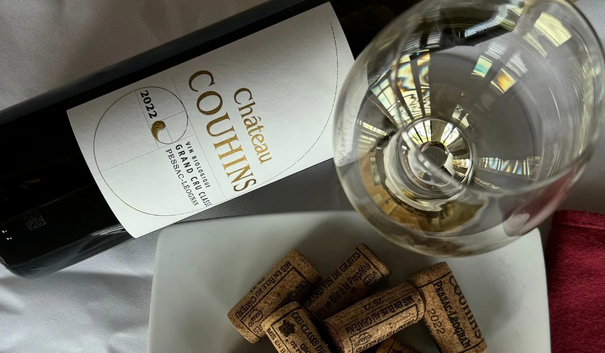 Bottle of Château Couhins 2022 white wine from Bordeaux with its label, accompanied by a filled glass and wine corks on a white plate