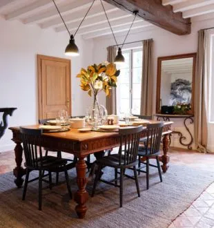 Elegant dining room featuring a solid wood table, black chairs, yellow flowers in a vase, exposed beams, and large windows overlooking the garden