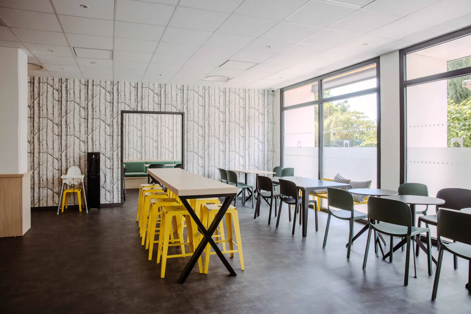 Modern coworking space featuring high tables with yellow chairs, green sofa, large windows overlooking a terrace, tree-patterned wallpaper, and suspended white ceiling.