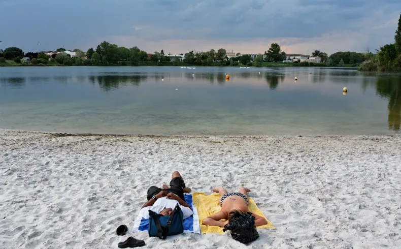 Two people relaxing on the sand at Bègles Plage in Bordeaux, overlooking a calm lake with residential buildings in the background