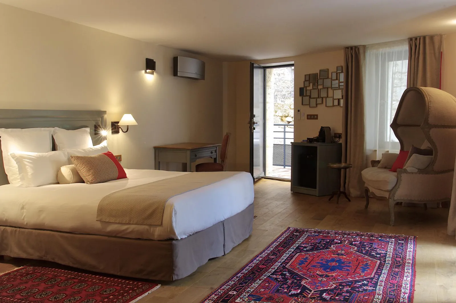 Elegant hotel room featuring a king-size bed, beige and wood furnishings, comfortable armchair, desk, private balcony, and oriental patterned red carpet at Hotel Vatel in Bordeaux.