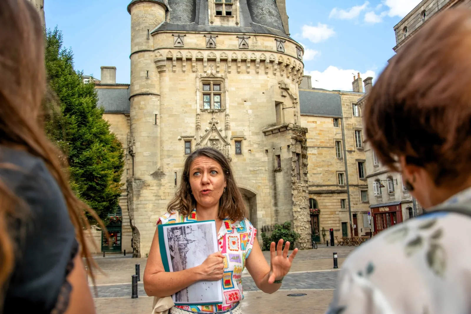 Tour guide holding an architectural plan in front of the Lantern Tower in Bordeaux, addressing a group of visitors in a paved courtyard.