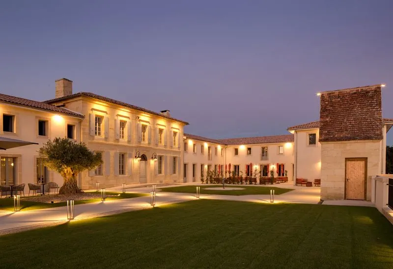 Night view of Château Fage in Bordeaux: elegant classical manor with light-colored stone facades, illuminated by spotlights, surrounding an inner courtyard with green lawn and paved pathways.