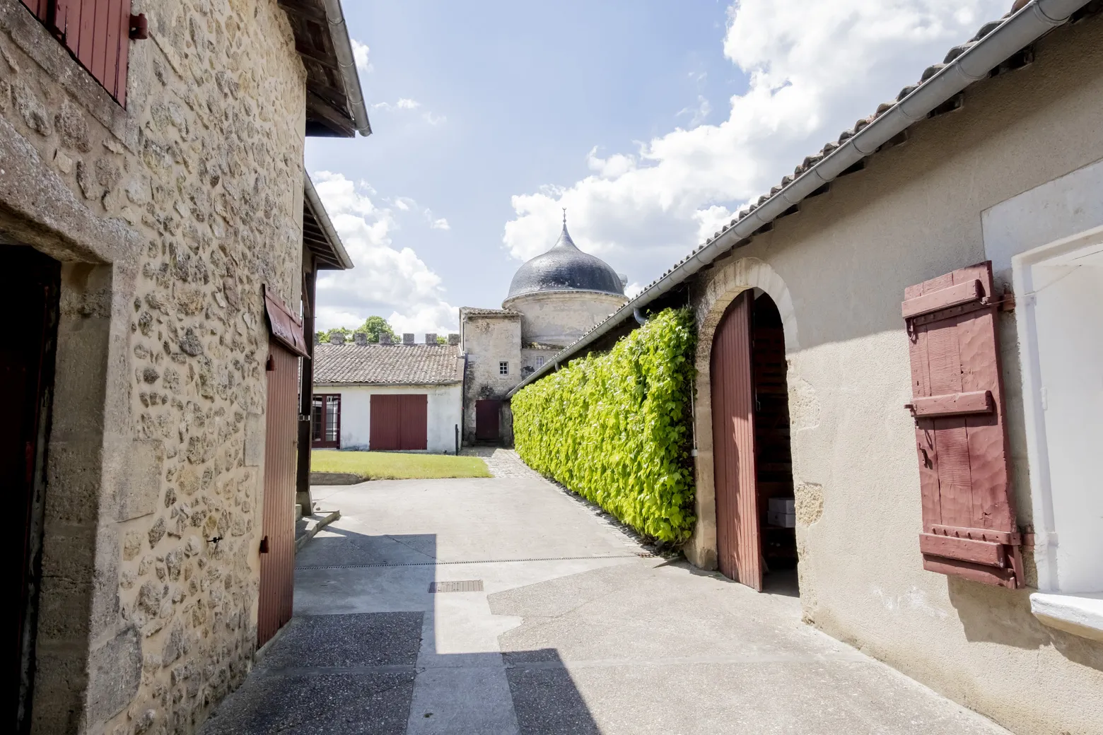 Inner courtyard of Château de Malle in Bordeaux featuring stone buildings, red doors, green hedge, and blue dome in the background